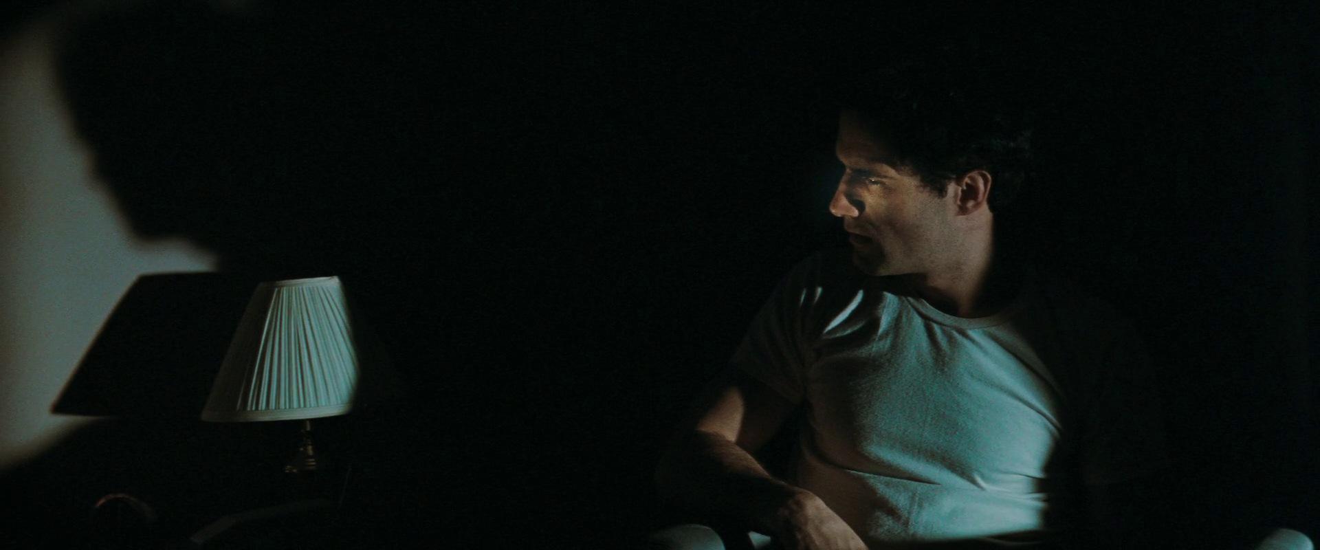 A man in a white t-shirt sits in bed in the dark
