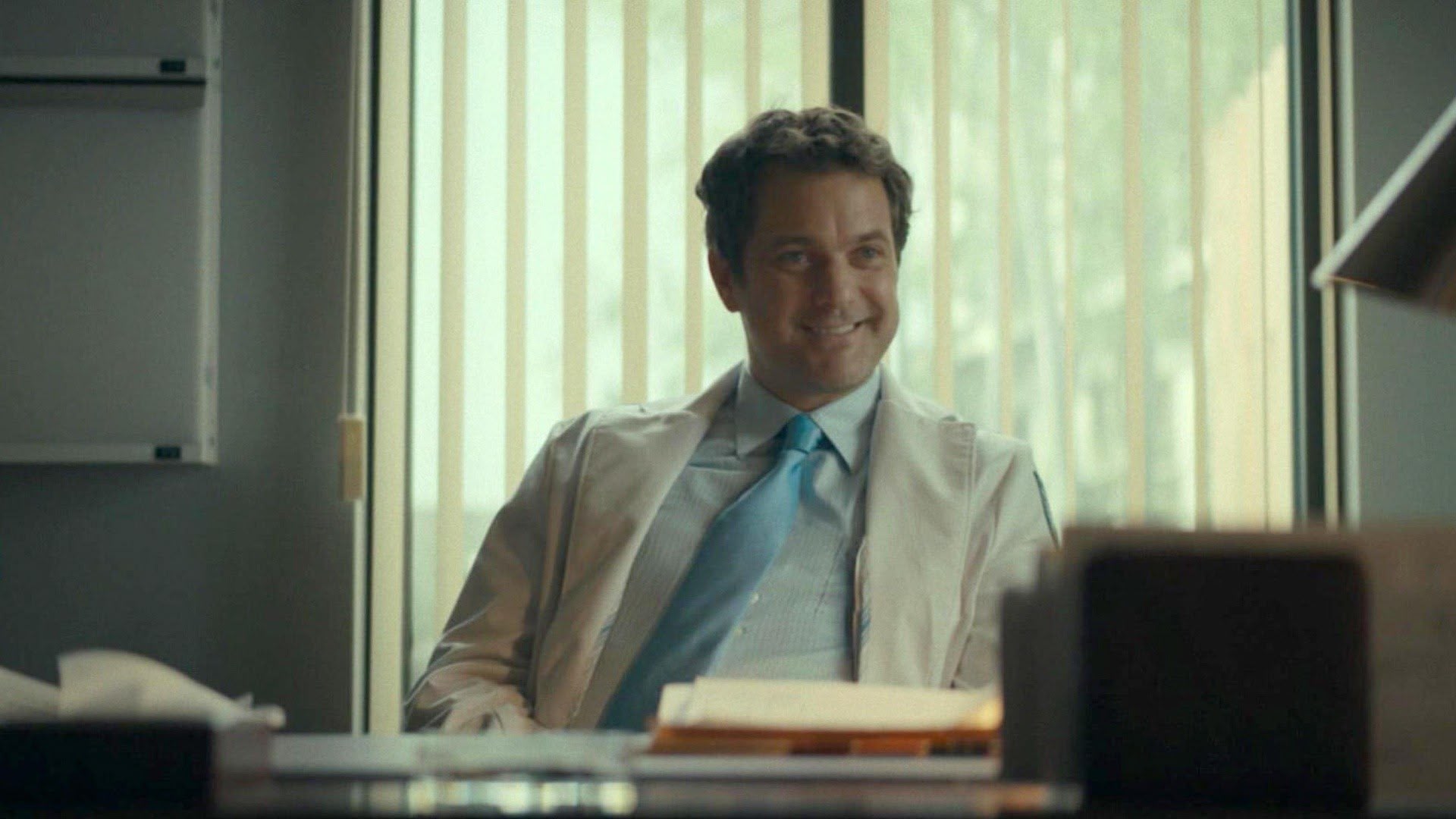 A smiling dark haired man wearing a light blue tie, a white dress shirt, and a doctor's white coat, seated behind a desk