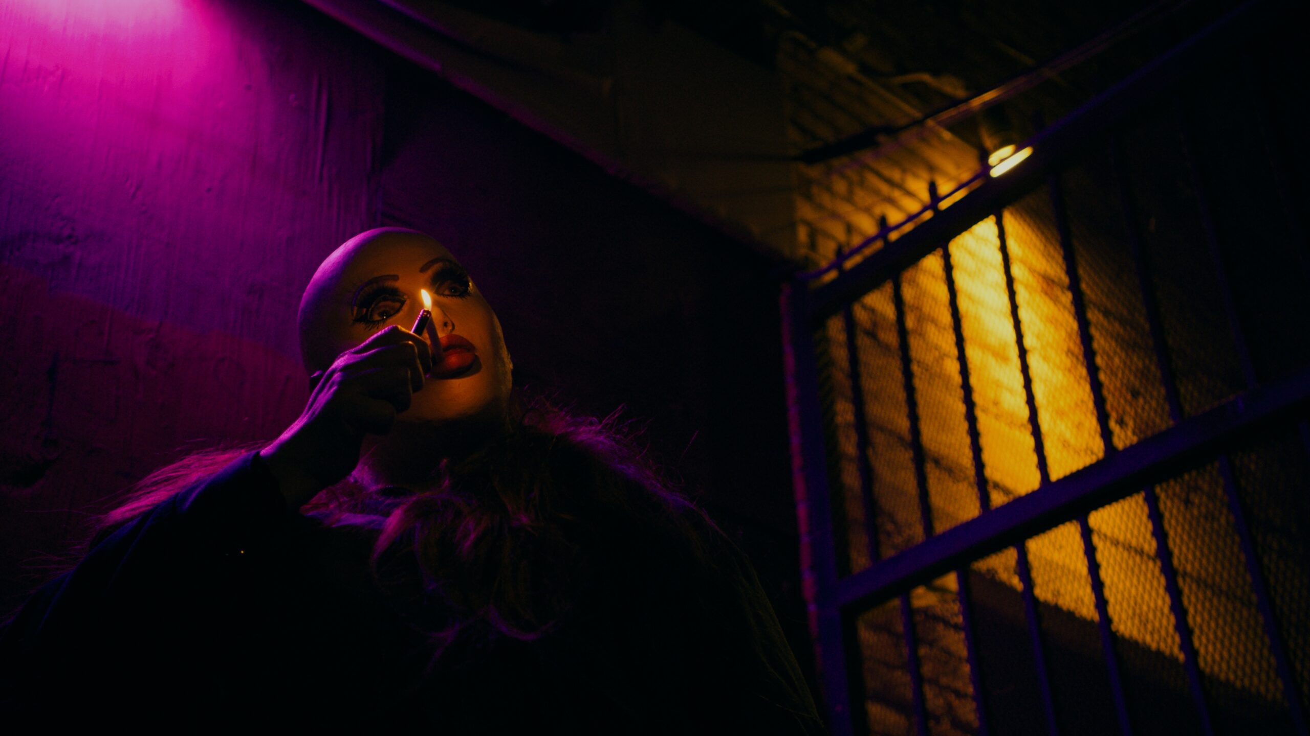 Frankie Maniac Woman sitting in dark room backlit in purple and gold, smoking a cigaretty through a luchador mask